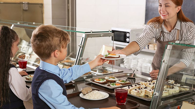 School cafeteria employee handing out egg and hash brown breakfast to a young school boy.